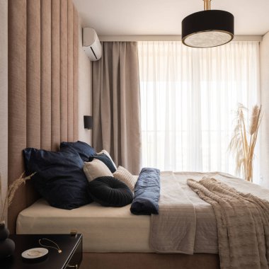 Dekoratif perdelerin arkasındaki büyük pencereli bej renkli şık bir yatak odası ve şık çarşaflı rahat bir yatak.