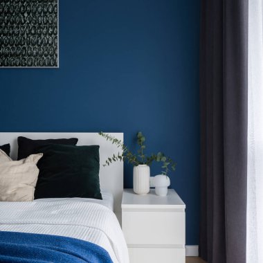 Yatak odasındaki rahat yatağın yanında koyu mavi duvarı olan basit, beyaz komodinin üzerine yakın plan.