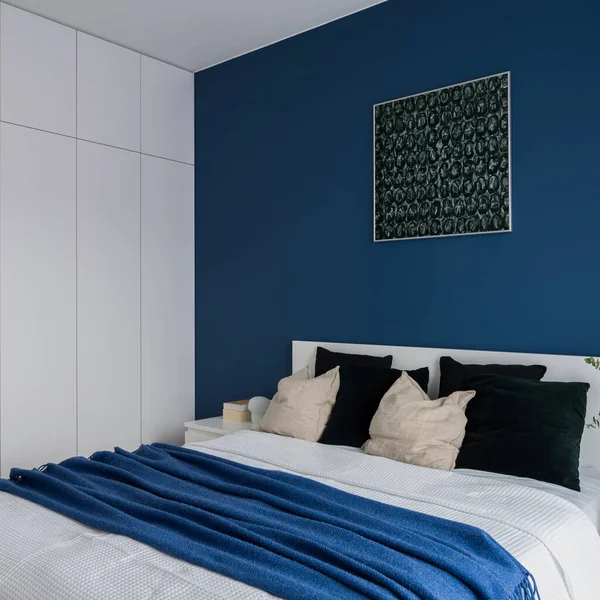 Cozy bed with simple bedclothes in modern bedroom with big white, wardrobe and stylish blue wall with art