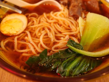 Asian noodle soup with braised beef, soft-boiled egg, bok choy, and yellow noodles in rich broth. Served in patterned bowl with chopsticks and spoon on rustic wooden table.