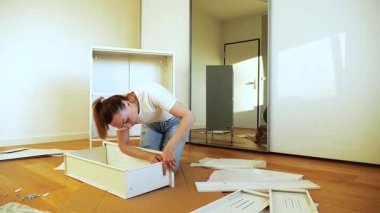 Genç bir kadın evde beyaz düz paket mobilya monte ediyor, tahta zeminde aletler ve vidalarla panelin üzerine yaslanıyor. DIY ev geliştirme eğitmeni, adım adım mobilya kurulum süreci