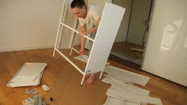 Genç bir kadın evde beyaz düz paket mobilya monte ediyor, tahta zeminde aletler ve vidalarla panelin üzerine yaslanıyor. DIY ev geliştirme eğitmeni, adım adım mobilya kurulum süreci