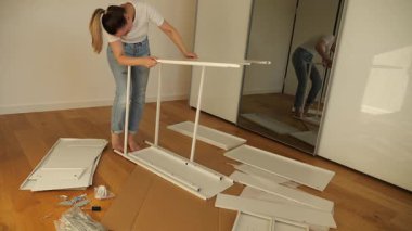 Genç bir kadın evde beyaz düz paket mobilya monte ediyor, tahta zeminde aletler ve vidalarla panelin üzerine yaslanıyor. DIY ev geliştirme eğitmeni, adım adım mobilya kurulum süreci