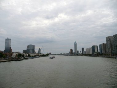 Rotterdam 'ın modern silueti Erasmus Köprüsü, nehirdeki tekneler ve bulutlu gökyüzünün altındaki yüksek binalar. Kentsel mimari, bu ikonik Hollanda şehrinde su yolu taşımacılığıyla buluşur.