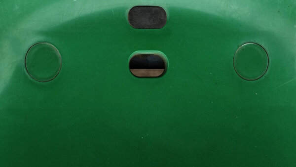 Green plastic chair backrest with two round indentations and two rectangular slots. Scratched surface, black handle detail, symmetrical design, and signs of everyday use.