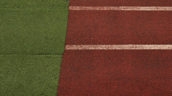 Крупный план зеленой искусственной территории, пересекающейся с красной беговой дорожкой с белой полосой. Контраст поверхности спортивного поля для легкой атлетики, футбола или тренировочных визуальных эффектов. Модульная трава и текстурированный путь.