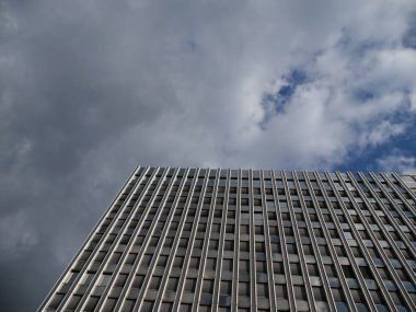 Şebeke görünümlü, üniformalı pencereli, aşağıdan vurulmuş, uzun bir ofis binası. Geometrik mimari şehir manzarası, gayrimenkul veya editoryal görseller için bulutlu gökyüzü ile buluşur.