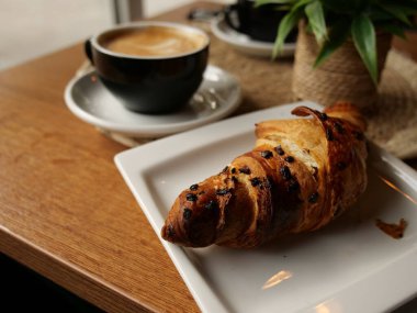 Ahşap masada kruvasan ve cappuccino ile sıcak kafe kahvaltı sahnesi. Doğal ışık, saksı bitkisi ve sıcak tonlarda rahat, davetkar bir ortam yaratır. Yaşam tarzı ya da editör kullanımı için..