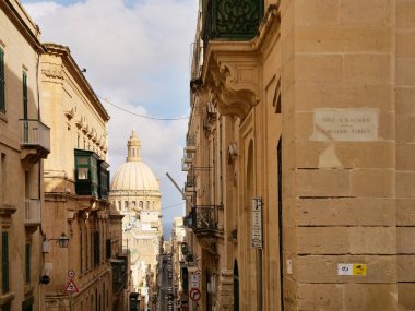 Malta Valletta 'da klasik bir sokak manzarası. St. Paul Katedrali' nin kubbesine doğru dar bir sokağa bakıyor. Fotoğraf, tarihi altın kireçtaşı mimarisini, geleneksel kapalı balkonları ve Wetstreet 'te birkaç aracı yakalıyor.,