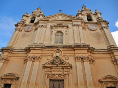 Malta 'nın antik müstahkem şehri Mdina' daki St. Paul Katedrali 'nin girişi. Görüntü, barok kireç taşı mimarisi, geniş sütunlar ve ana kapının üstündeki parlak mavi gökyüzünün altında parlayan bulutlarla birlikte armanın altını çiziyor..