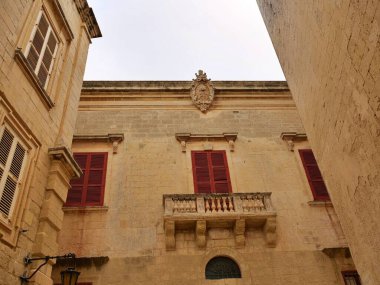 Geleneksel Malta kireç taşı ön cephesinde süslü bir barok taş balkon ve parçalı bulutlu bir gökyüzünün altında kapalı pencere panjurları. Bu Mdina, Malta gibi tarihi şehirlerde bulunan klasik Akdeniz mimarisidir..
