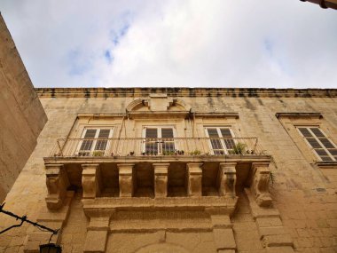 Geleneksel Malta kireç taşı ön cephesinde süslü bir barok taş balkon ve parçalı bulutlu bir gökyüzünün altında kapalı pencere panjurları. Bu Mdina, Malta gibi tarihi şehirlerde bulunan klasik Akdeniz mimarisidir..