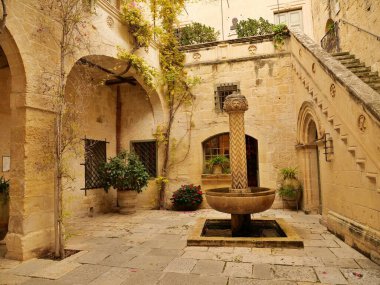 Malta 'nın Mdina kentindeki bir ortaçağ müzesi olan Palazzo Falson' ın sakin, güneşli merkezi avlusunun dikey görüntüsü. Sahnede yaşlanmış, altın renkli kireç taşı mimarisi, Rönesans tarzı merdiven, kemerli kapılar, saksı bitkileri yer alıyor.,