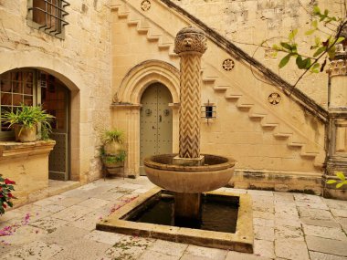 Malta 'nın Mdina kentindeki bir ortaçağ müzesi olan Palazzo Falson' ın sakin, güneşli merkezi avlusunun dikey görüntüsü. Sahnede yaşlanmış, altın renkli kireç taşı mimarisi, Rönesans tarzı merdiven, kemerli kapılar, saksı bitkileri yer alıyor.,