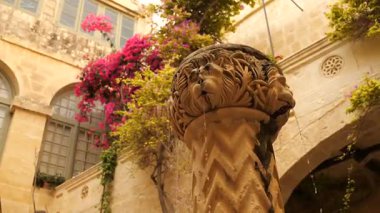 Mdina, Malta 'da bulunan Palazzo Falson Tarih Müzesi bahçesinde, fıskiyeler, merdivenler üzerinde aslan heykeli ve parlak, güneşli bir gökyüzü altında geleneksel kireç taşı mimarisi yer alıyor..