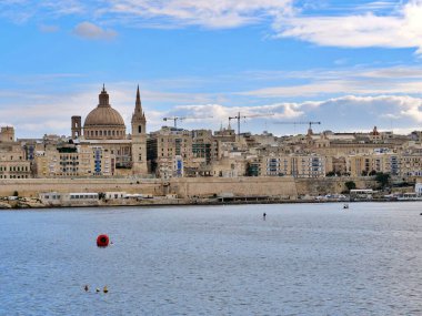 Limandan tarihi Valletta siluetinin panoramik manzarasında, Carmel Dağı 'nın Leydisi Bazilikası' nın ikonik evi ve güçlendirilmiş kireçtaşı şehir duvarlarının üzerinde yükselen Aziz Paul Katedrali 'nin kulesi yer alıyor..