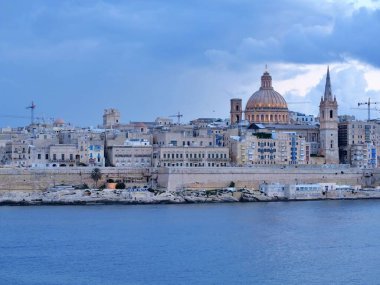 Limandan tarihi Valletta siluetinin panoramik manzarasında, Carmel Dağı 'nın Leydisi Bazilikası' nın ikonik evi ve güçlendirilmiş kireçtaşı şehir duvarlarının üzerinde yükselen Aziz Paul Katedrali 'nin kulesi yer alıyor..