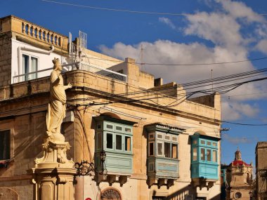 Geleneksel Malta mimarisi süslenmiş mavi ahşap balkonlar (gallariji), bir köşede önde gelen bir dini heykel, ve tarihi kireçtaşı duvar Mdina, Malta 'da güneşli bir şehir sokak sahnesi.