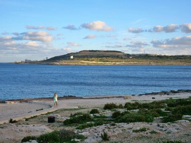 Elbiseli kadın, kıyı yolunda, kayalar ve bitkilerle güneşli gökyüzünün altındaki mavi okyanusa doğru yürüyor.