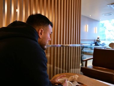 Modern bir kafede oturan sakallı genç bir adamın yan profili. Kirli sakallı, siyah kapüşonlu düşünceli bir adam ahşap kaplama arka planlı kapalı bir ortama bakıyor. Elinde bir fincan kahve ya da çay olan bir adamın özgün yaşam tarzı portresi.