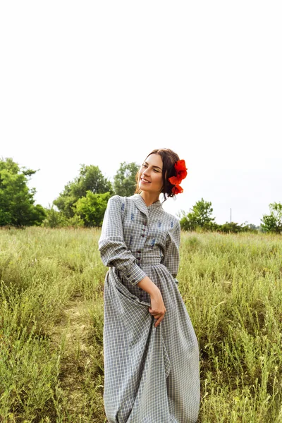 Outdoor portret van een mooie brunette vrouw in jurk met rode bloemen in haar haar in het veld. — Stockfoto