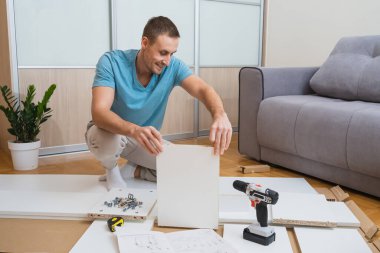 Mavi tişörtlü gülümseyen adam yerde diz çöker ve parlak bir odada bir tornavida ve talimatlarla mobilya parçaları birleştirir. Yüksek kalite fotoğraf