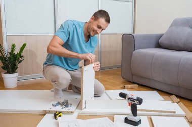 Mavi tişörtlü gülümseyen adam yerde diz çöker ve parlak bir odada bir tornavida ve talimatlarla mobilya parçaları birleştirir. Yüksek kalite fotoğraf