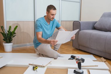 Adam yerde oturup mobilya montaj talimatlarını okuyor. Aletler ve düz paket parçaları etrafına saçılmış, aydınlık ve modern bir oturma odasında.