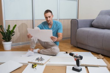 Adam yerde oturup mobilya montaj talimatlarını okuyor. Aletler ve düz paket parçaları etrafına saçılmış, aydınlık ve modern bir oturma odasında.