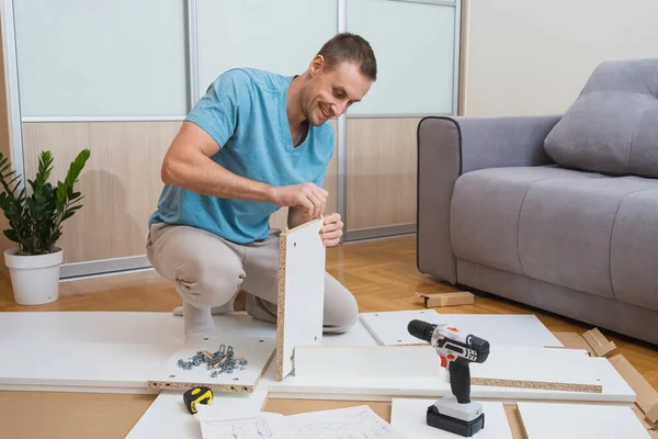 Mavi tişörtlü gülümseyen adam yerde diz çöker ve parlak bir odada bir tornavida ve talimatlarla mobilya parçaları birleştirir. Yüksek kalite fotoğraf