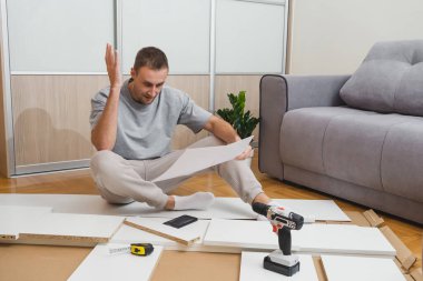 Dağınık parçalar ve aletler arasındaki mobilya talimatlarını okurken kafası karışmış bir adam, parlak modern oda DIY sorun çıkaran bir nalbur dükkanında ev yenileme blogları yazıyor. Yüksek kalite fotoğraf