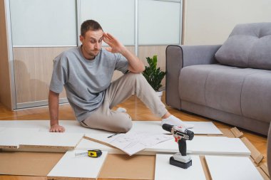 Dağınık parçalar ve aletler arasındaki mobilya talimatlarını okurken kafası karışmış bir adam, parlak modern oda DIY sorun çıkaran bir nalbur dükkanında ev yenileme blogları yazıyor. Yüksek kalite fotoğraf