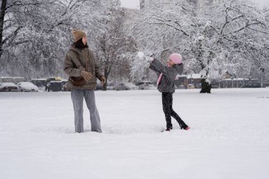 Pembe şapkalı gülümseyen bir kız karlı kış parkında gülen annesine kartopu atıyor. Arka planda ağaçlar ve arabalar var. Eğlenceli kış aktiviteleri..