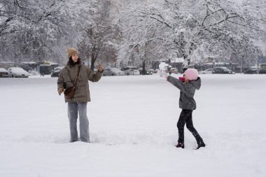 Pembe şapkalı gülümseyen bir kız karlı kış parkında gülen annesine kartopu atıyor. Arka planda ağaçlar ve arabalar var. Eğlenceli kış aktiviteleri..