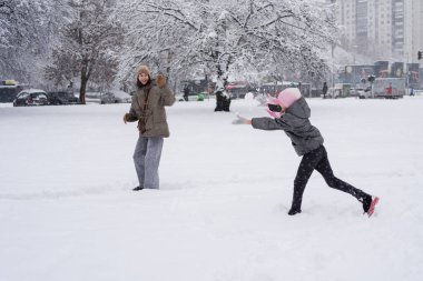 Pembe şapkalı gülümseyen bir kız karlı kış parkında gülen annesine bir kartopu atar. Arka planda ağaçlar ve arabalar, neşeli açık hava aktiviteleri aile eğlence gezisi reklamları..