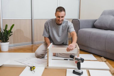 Gülümseyen adam yerde diz çöker ve parlak bir odada bir tornavida ve talimatlarla mobilya parçaları birleştirir. Yüksek kalite fotoğraf