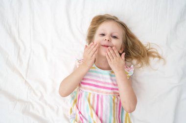 Yatağa uzanmış bir kız, elleri yüzüne yakın bir şekilde öpüşür ve neşeli, neşeli bir çocukluk anı aile hayatı reklamında geniş bir ağız açar. Yüksek kalite fotoğraf