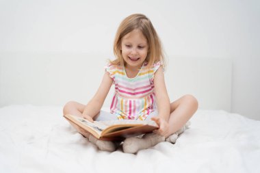 Gülümseyen bir kız yatağında bağdaş kurup iki eliyle büyük bir kitap okuyarak minimalist bir odada rahat rahat çocukluk edebiyat eğitimi aile yaşam tarzı kavramını öğreniyor. Yüksek kalite fotoğraf
