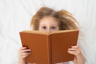 Geniş, meraklı gözleriyle geniş bir kitabın tepesinde yatağında uzanan bir kız. Rahat, aydınlık bir odada çocukluğunda hayal gücü aile hayatı reklamları okuyordu. Yüksek kalite fotoğraf