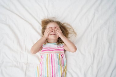 Parlak beyaz bir odada, renkli çizgili pijama giyen neşeli çocukluk aile hayatı konseptini takınıp, elleri hafifçe gülümseyerek yatağa uzanan bir kız gözlerini kapatır. Yüksek kalite fotoğraf