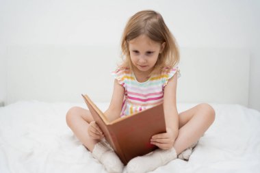 Gülümseyen bir kız yatağında bağdaş kurup iki eliyle büyük bir kitap okuyarak minimalist bir odada rahat rahat çocukluk edebiyat eğitimi aile yaşam tarzı kavramını öğreniyor. Yüksek kalite fotoğraf