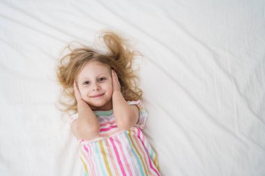 Yatakta uzanmış gülümseyen bir kız, elleri parlak beyaz bir odada, renkli çizgili pijamalarıyla kulaklarını kapatıyor. Neşeli çocukluk anı aile hayatı konsepti. Yüksek kalite fotoğraf