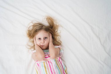 Yatakta uzanan bir kız, elleri parlak beyaz bir odada, renkli çizgili pijamalar giyerek kulaklarını kapatıyor ve gürültülü bir çocukluk sesi duymamış gibi davranıyor. Yüksek kalite fotoğraf