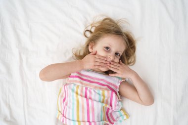 Yatağa uzanmış bir kız, elleri yüzüne yakın bir şekilde öpüşür ve neşeli, neşeli bir çocukluk anı aile hayatı reklamında geniş bir ağız açar. Yüksek kalite fotoğraf