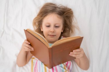 Sırtüstü uzanmış bir kız, geniş bir kitap okur. Odaklanmış, parlak, rahat bir odada. Yukardan aşağı bakan bir evde çocukluk dönemi okur yazarlık aile hayatı konsepti reklamları okuyan bir kız. Yüksek kalite fotoğraf