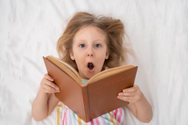 Yatakta ağzı açık bir şekilde uzanan bir kız, açık bir kitap tutar. Açık çizgili, yumuşak, ılık, ev yapımı rahat giysileri olan, hayal gücü ve aile yaşam tarzı kavramları okuyan bir kız. Yüksek kalite fotoğraf