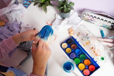 Bir çocuğun elleri boya bardağı ve paleti olan bir masanın üzerine fırçayla oyuncak bebeklerin saçlarını maviye boyar. Etrafı bitkilerle ve sanat malzemeleriyle çevrilidir. Yüksek kalite fotoğraf