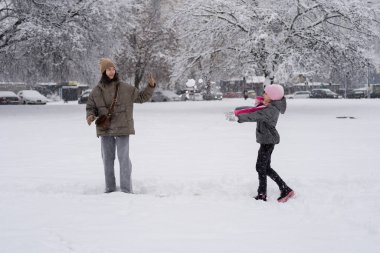 Pembe şapkalı gülümseyen bir kız karlı kış parkında gülen annesine kartopu atıyor. Arka planda ağaçlar ve arabalar var. Eğlenceli kış aktiviteleri..