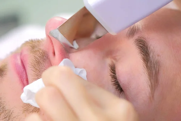 Cosmetologist makes ultrasonic face cleaning procedure to young man in clinic.