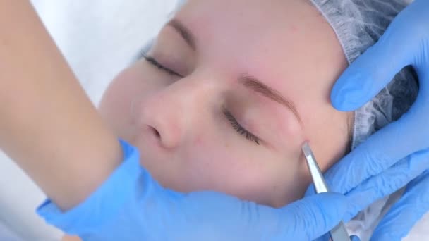 Cosmetologist plucks out hair on eyebrows with tweezers for woman, face closeup. — Stock Video